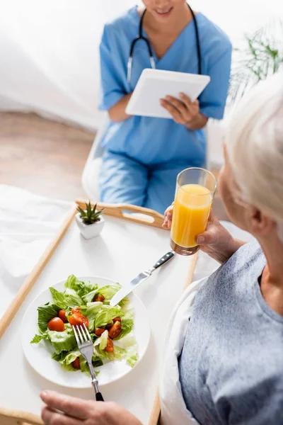 Vista ad alto angolo della donna anziana che fa colazione vicino all'infermiera utilizzando tablet digitale su sfondo sfocato — Foto stock