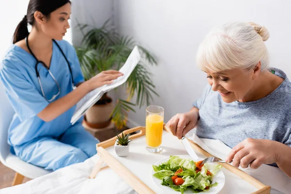 Piacere anziana donna avendo colazione vicino giovane asiatico infermiera lettura giornale su sfondo sfocato — Foto stock