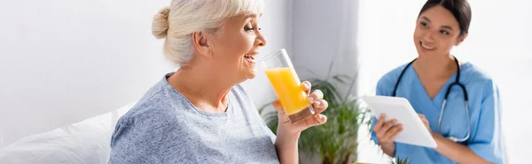 Радісна старша жінка п'є апельсиновий сік біля усміхненої азіатської медсестри з цифровим планшетом на розмитому фоні, банер — стокове фото