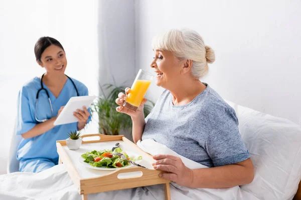 Zufriedene ältere Frau frühstückt und trinkt Orangensaft neben lächelnder asiatischer Krankenschwester auf verschwommenem Hintergrund — Stockfoto