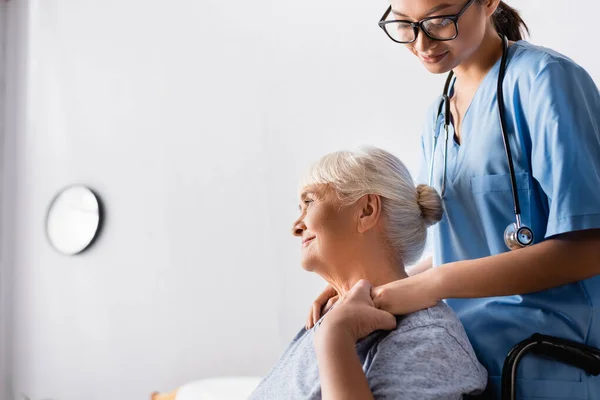 Sorrindo sênior mulher segurando mão de jovem asiático enfermeira enquanto olhando afastado no hospital — Fotografia de Stock