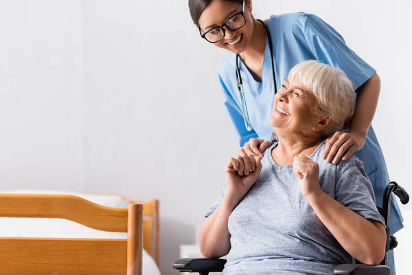 Happy asian nurse touching shoulders of aged handicapped woman laughing in wheelchair — Stock Photo