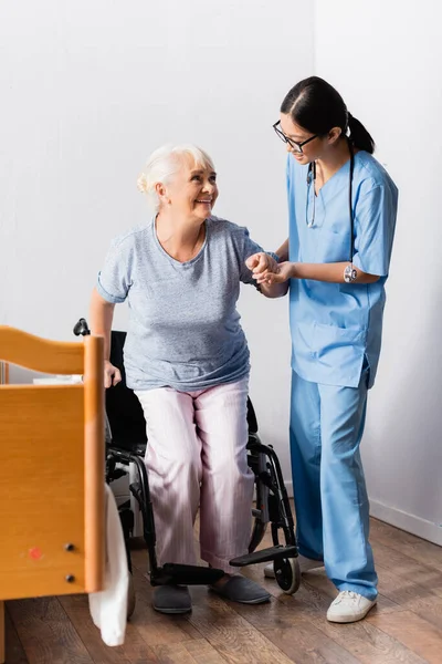 Молодая азиатская медсестра помогает счастливой пожилой женщине встать с инвалидного кресла — стоковое фото
