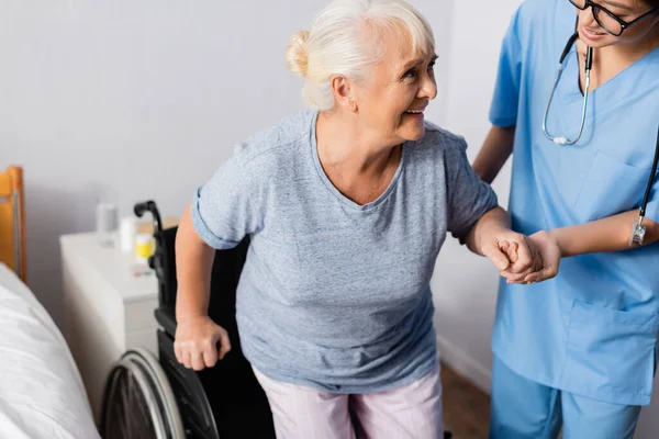 Asiatische Krankenschwester unterstützt positive Seniorin beim Aufstehen aus dem Rollstuhl — Stockfoto