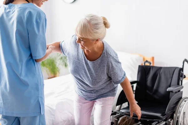 Enfermera joven ayudando a una mujer anciana a levantarse de la silla de ruedas - foto de stock