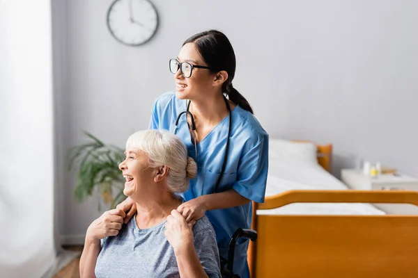 Ältere, lachende behinderte Frau hält die Hände einer jungen asiatischen Krankenschwester — Stockfoto
