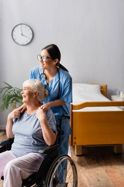 Cheerful, senior disabled woman laughing while touching hands of young asian nurse — Stock Photo