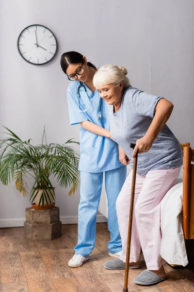 Mujer asiática ayudando a una mujer anciana con bastón de caminar levantándose de la cama en el hospital - foto de stock