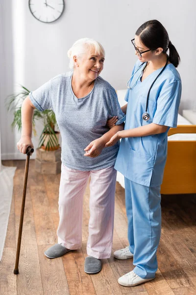 Elderly woman with walking stick looking at young asian nurse supporting her in hospital — Stock Photo