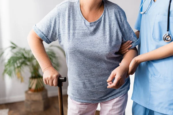 Partial view of nurse supporting elderly woman with walking stick in hospital — Stock Photo