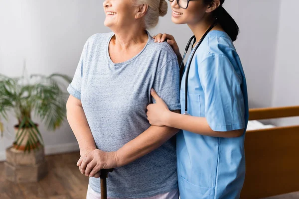 Vista recortada de la enfermera sonriente que apoya a la mujer anciana positiva con bastón en el hospital - foto de stock