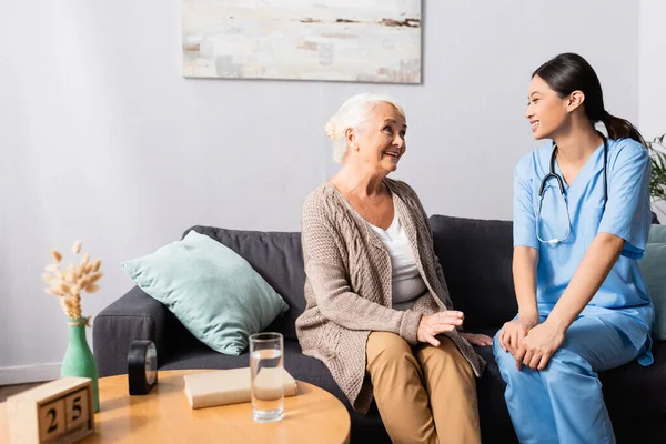 Sorridente infermiera asiatica e allegra donna anziana che parla sul divano in casa di cura, in primo piano sfocato — Foto stock