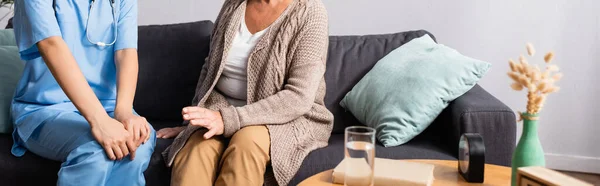 Ausgeschnittene Ansicht von Seniorin und Krankenschwester auf Sofa im Pflegeheim sitzend, verschwommener Vordergrund, Banner — Stockfoto