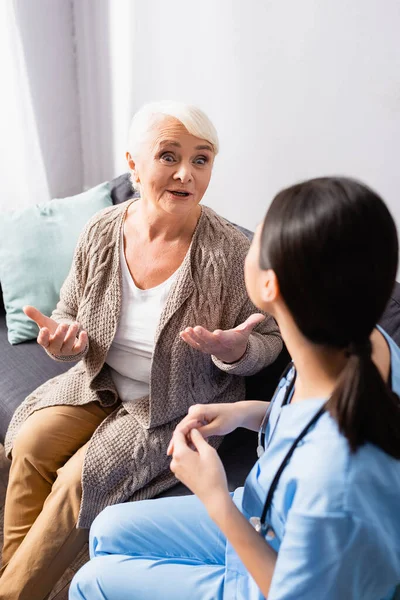 Изумленная пожилая женщина жестом во время разговора с медсестрой в доме престарелых, размытый передний план — стоковое фото