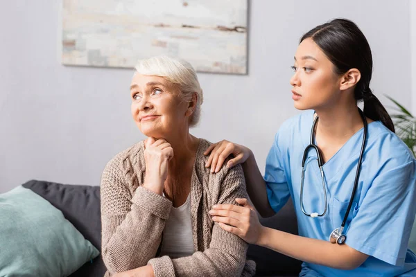 Молодая азиатская медсестра успокаивает расстроенную пожилую женщину сидя и глядя в дом престарелых — стоковое фото