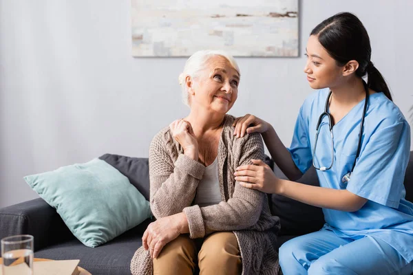 Young asian nurse touching smiling senior woman during conversation in nursing home — Stock Photo