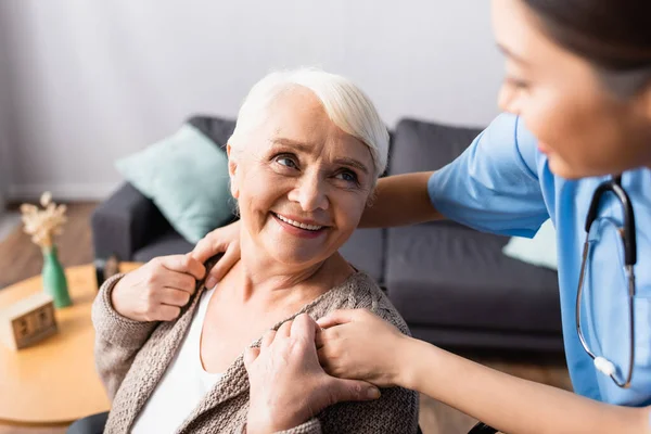Happy elderly woman touching hands of young asian nurse on blurred foreground — Stock Photo