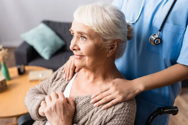 Enfermera tocando hombros de mujer discapacitada soñadora sonriendo mientras mira hacia otro lado - foto de stock