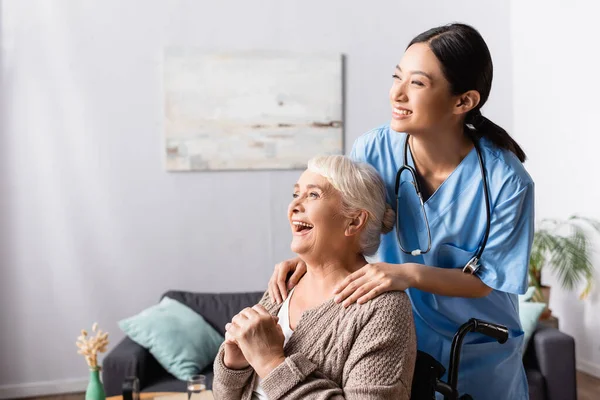 Щаслива азіатська медсестра торкається плечей літньої інвалідної жінки, яка сміється в інвалідному візку — стокове фото