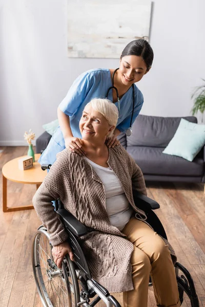 Sorridente infermiera asiatica toccare le mani di allegra donna anziana seduta in sedia a rotelle — Foto stock