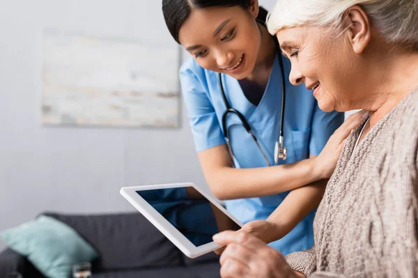 Sonriente asiático enfermera holding digital tablet con blanco pantalla cerca feliz anciano mujer, borrosa fondo - foto de stock