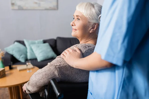 Nurse touching shoulder of aged, smiling disabled woman in wheelchair, blurred foreground — Stock Photo