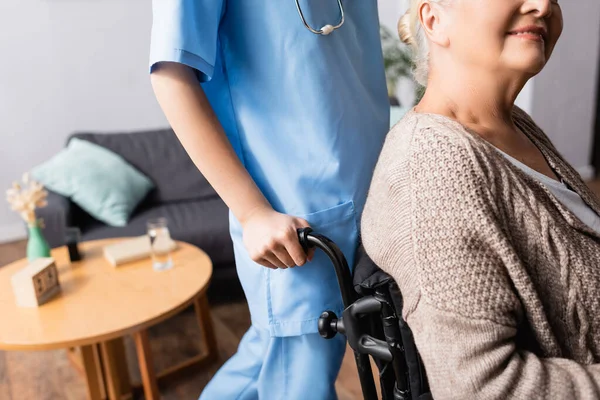Partial view of nurse near smiling handicapped woman sitting in wheelchair, blurred background — Stock Photo