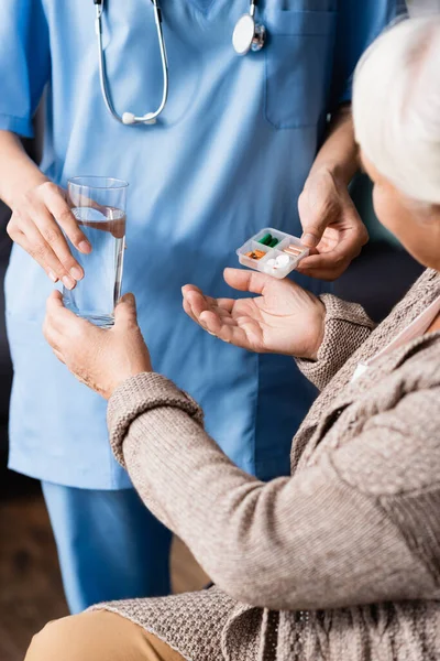 Vista ritagliata di infermiera dando pillole e bicchiere d'acqua alla donna anziana, sfondo sfocato — Foto stock