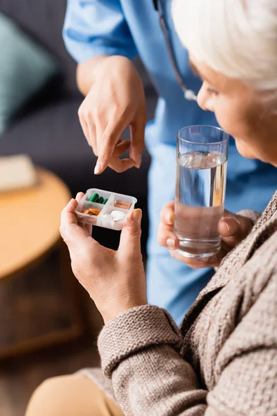 Infirmière pointant vers les pilules en main de la femme âgée avec un verre d'eau, fond flou — Photo de stock