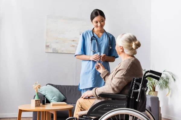 Smiling asian nurse holding glass of water near aged disabled woman in wheelchair — Stock Photo