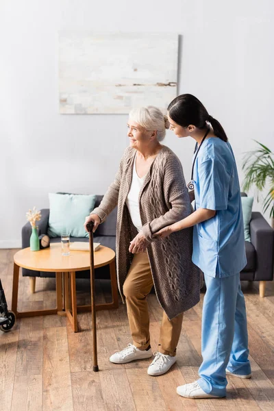 Young asian nurse supporting senior woman with walking stick in nursery home — Stock Photo