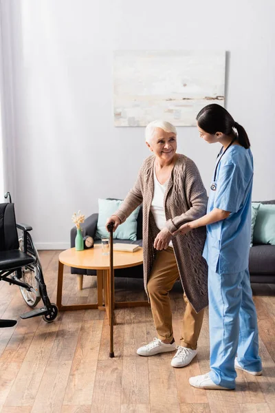 Positive elderly woman with walking stick looking at asian nurse supporting her in nursery home — Stock Photo