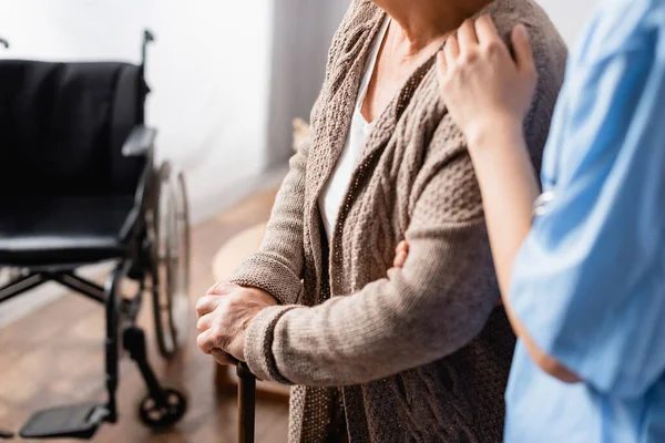 Vue recadrée de l'infirmière touchant l'épaule de la femme âgée avec bâton de marche près du fauteuil roulant sur fond flou — Photo de stock