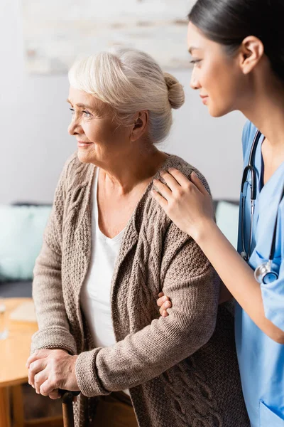 Young asian nurse supporting smiling elderly woman with walking stick — Stock Photo