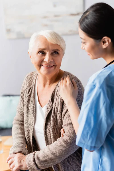 Junge asiatische Krankenschwester berührt Schulter einer lächelnden Seniorin im Pflegeheim, verschwommener Vordergrund — Stockfoto