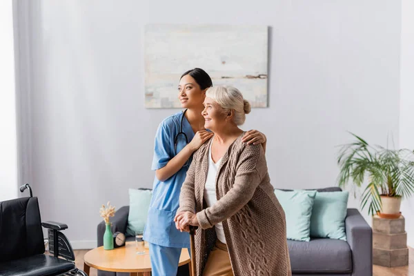 Young asian nurse hugging shoulders of aged smiling woman with walking stick in nursery home — Stock Photo