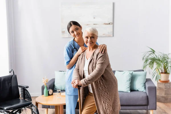 Smiling asian nurse and elderly woman with walking stick looking at camera in nursing home — Stock Photo