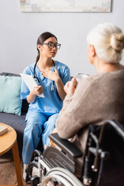 Young asian woman touching chest while talking with aged disabled woman sitting in wheelchair on blurred foreground — Stock Photo