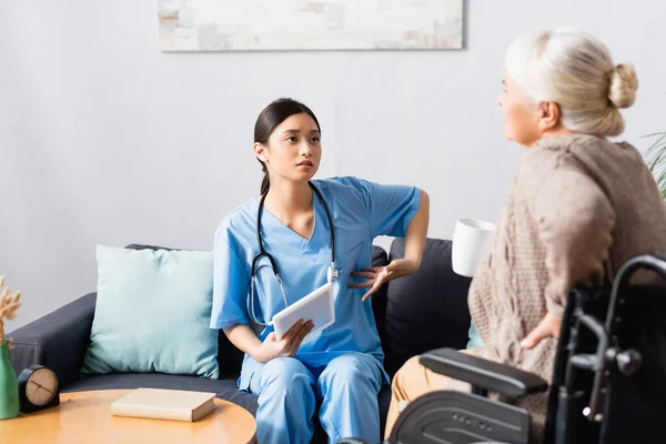 Asiatische Krankenschwester hält digitales Tablet in der Hand und zeigt mit der Hand auf sich selbst, während sie mit behinderter Frau im Rollstuhl im verschwommenen Vordergrund spricht — Stockfoto