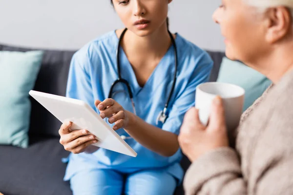 Cropped view of asian nurse using digital tablet near elderly woman holding cup of tea — Stock Photo