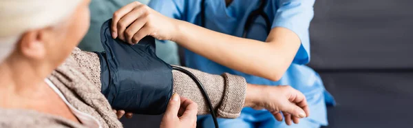 Partial view of nurse fixing cuff of tonometer on arm of senior woman on blurred foreground, banner — Stock Photo