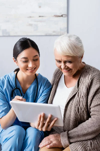 Fröhliche asiatische Krankenschwester zeigt mit dem Finger auf digitales Tablet neben lächelnder Seniorin — Stockfoto