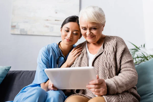 Happy asian woman leaning on shoulder of aged smiling woman holding digital tablet — Stock Photo