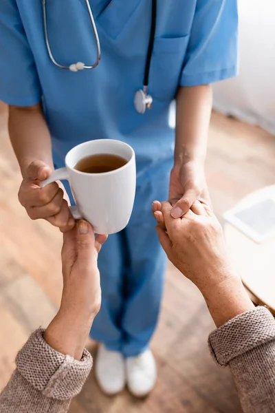 Vista ritagliata di infermiera dando tazza di tè alla donna anziana mentre tiene la mano, sfondo sfocato — Foto stock