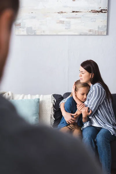 Depressed woman hugging kid on couch near husband on blurred foreground — Stock Photo
