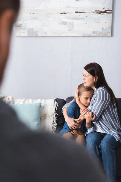 Depressive Frau umarmt Kind auf Couch neben Ehemann im verschwommenen Vordergrund — Stockfoto