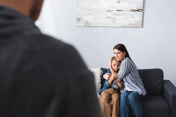 Frustrated woman embracing daughter on sofa near abusive husband on blurred foreground — Stock Photo