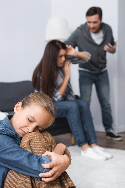 Оскорбленная девушка, сидящая рядом с жестоким отцом, ссорящаяся с матерью на размытом фоне — стоковое фото