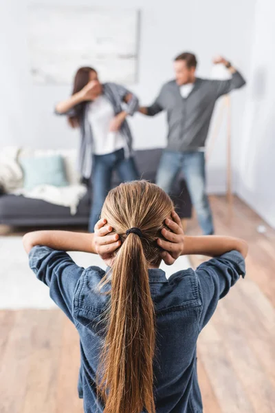 Daughter covering ears with hands while abusive father beating mother on blurred background at home — Stock Photo