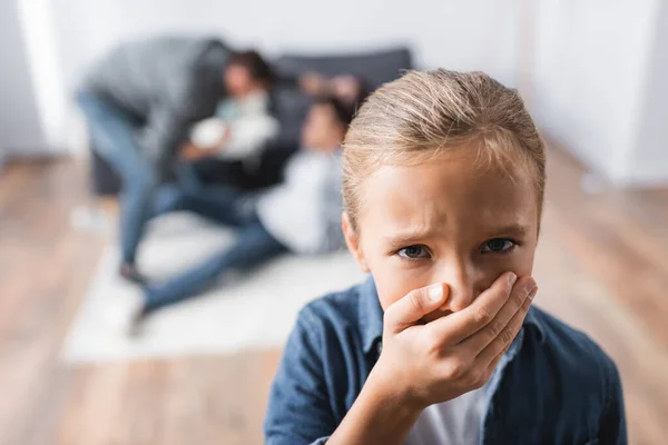 Niño ofendido cubriendo la boca con la mano mientras los padres pelean sobre un fondo borroso en la sala de estar - foto de stock